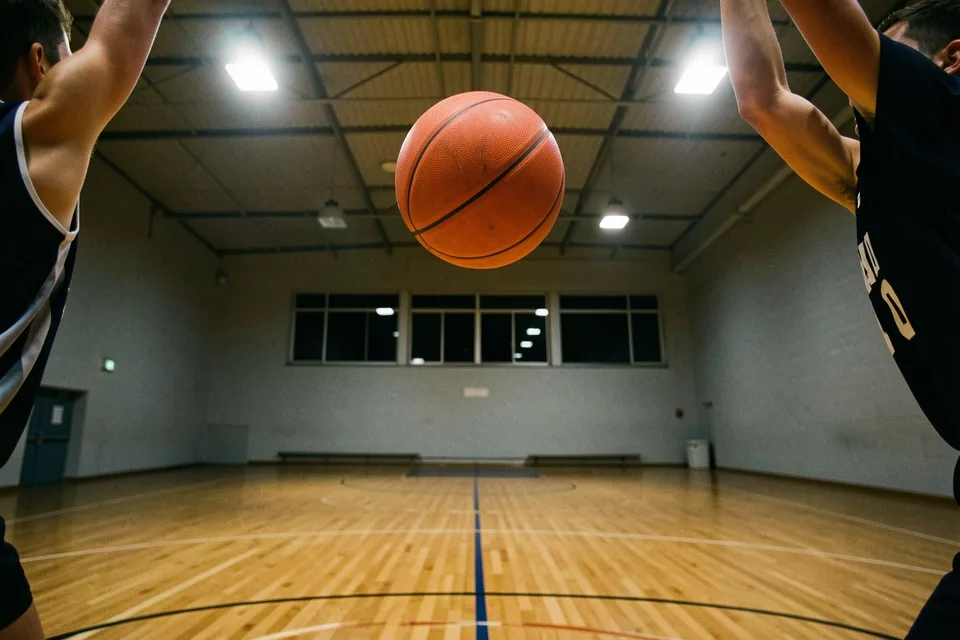 Η μπάλα στο ψηλότερο σημείο της τροχιάς της μέσα στο φωτισμένο δημοτικό γυμναστήριο, με δύο παίκτες στις άκρες του κάδρου.