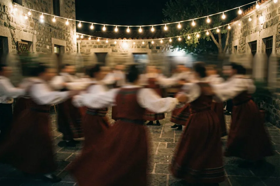 Χορευτές με παραδοσιακές κόκκινες φορεσιές στροβιλίζονται πιασμένοι χέρι χέρι κάτω από τα φωτάκια της αυλής.